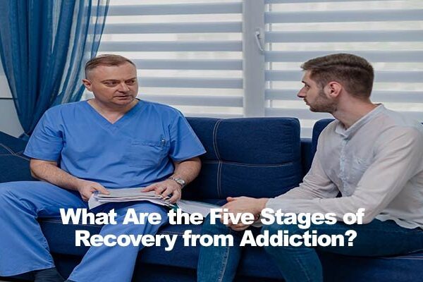 Two men sitting on a sofa discussing recovery, one in blue scrubs, the other in a white shirt.