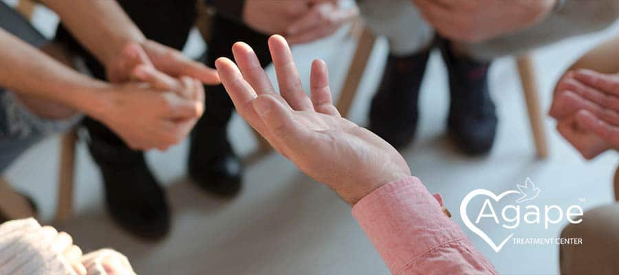 Person in a pink shirt gesturing with hands, surrounded by others sitting, Agape Treatment Center logo nearby.