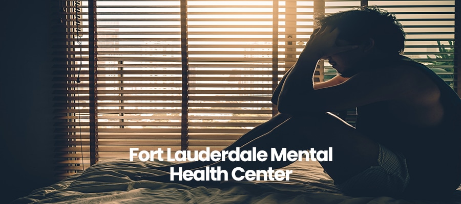 Person sitting on bed, holding head, with blinds in background. Text: Fort Lauderdale Mental Health Center.