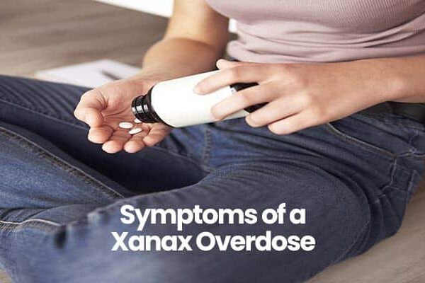 Person sitting on the floor in jeans holding pill bottle. Symptoms of a Xanax Overdose text overlay.