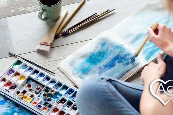 Person creating watercolor painting on wooden floor with paint palette and brushes nearby.