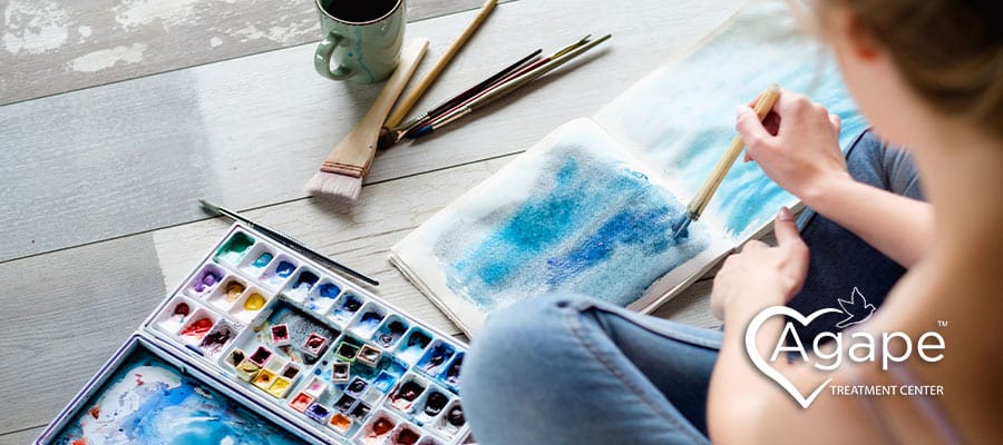 Person creating watercolor painting on wooden floor with paint palette and brushes nearby.