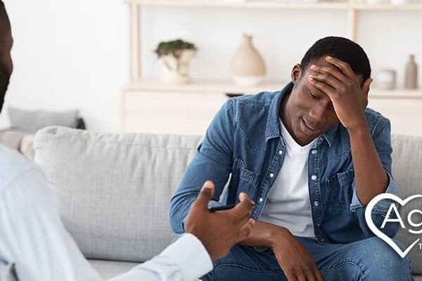 Person in denim shirt looking stressed with hand on head, sitting on couch.