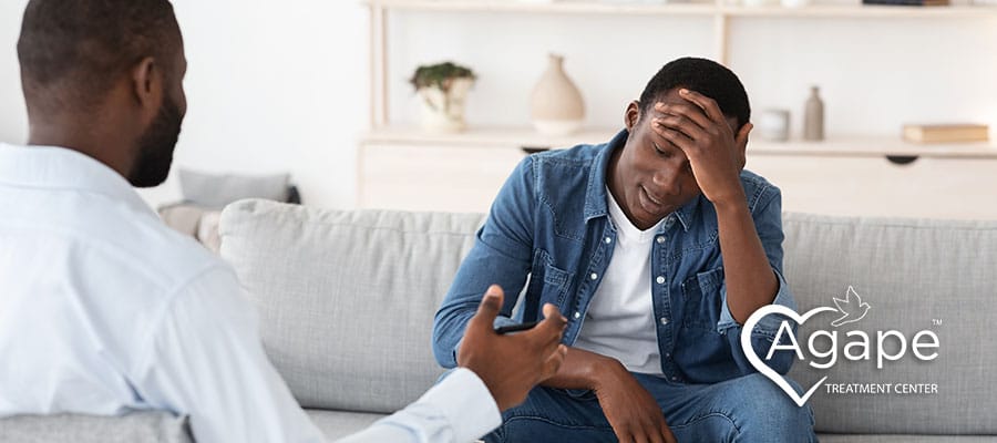 Person in denim shirt looking stressed with hand on head, sitting on couch.