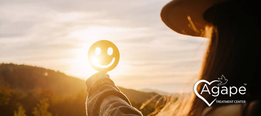 Person holding a smiling face cutout against the sunset with Agape Treatment Center logo.