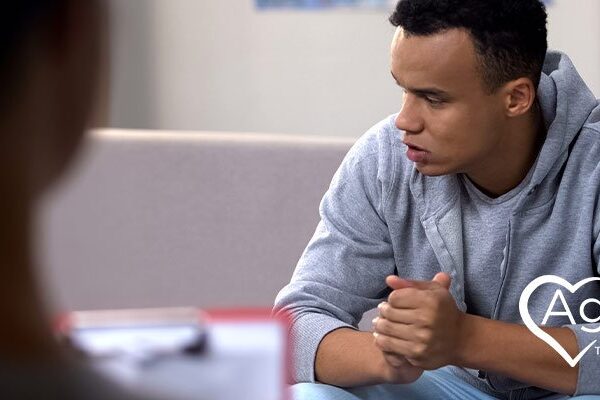 Person in gray hoodie sitting on couch with hands clasped.