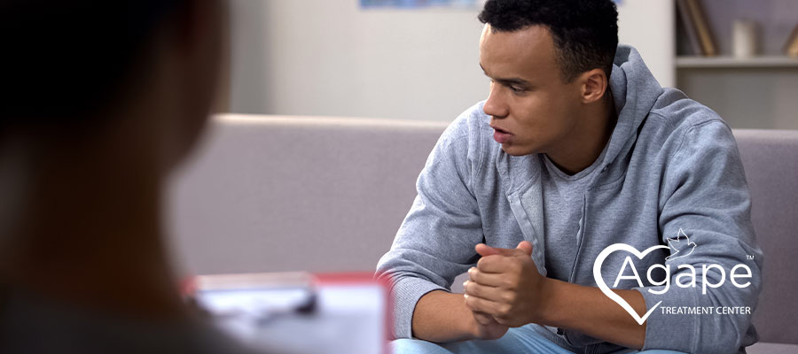 Person in gray hoodie sitting on couch with hands clasped.