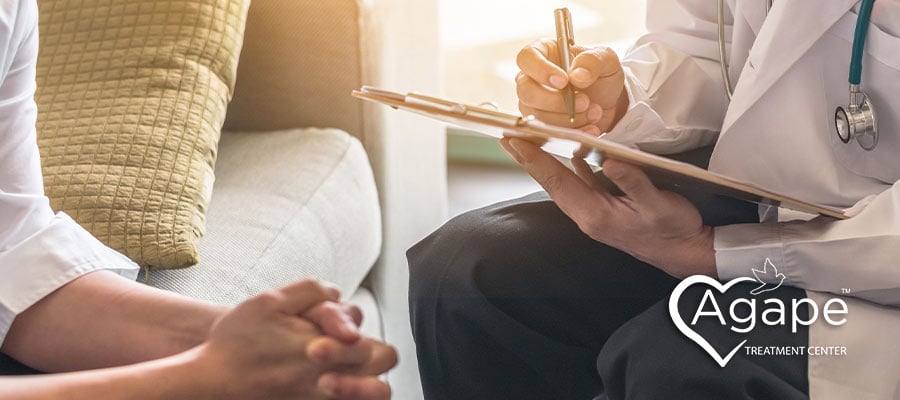 Person in white coat writing on clipboard, Agape Treatment Center logo.