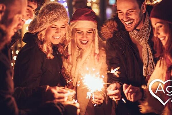 Group of people holding sparklers, wearing winter clothing, smiling under festive lights.