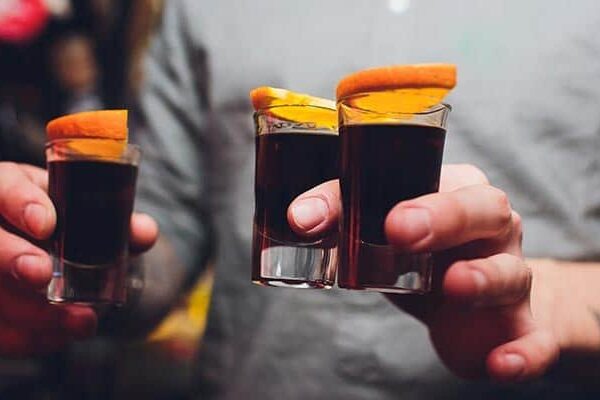 Person holding three shot glasses with orange slices.