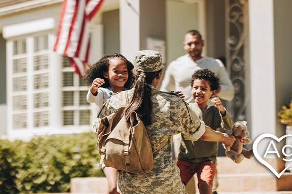 Person in military uniform hugging two children outside house.