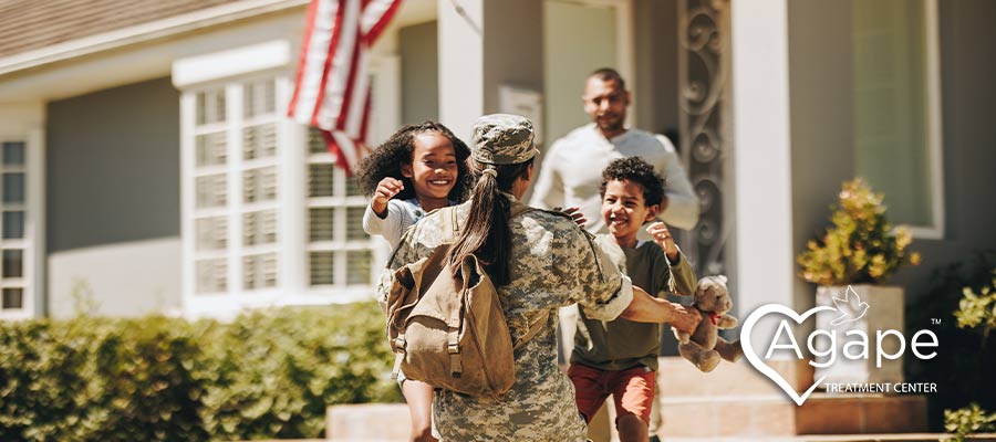 Person in military uniform hugging two children outside house.