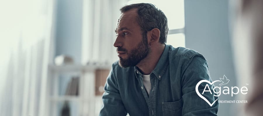 Person in denim shirt sitting and looking thoughtful with Agape Treatment Center logo.