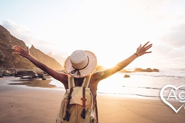 Person wearing a hat and backpack with arms raised on a beach at sunset.