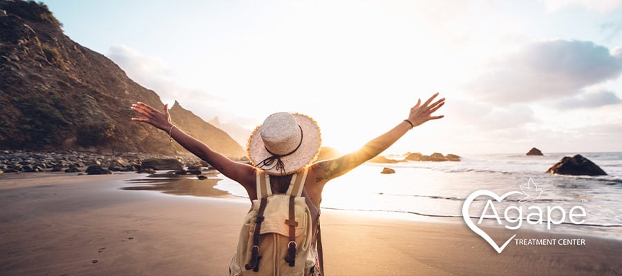 Person wearing a hat and backpack with arms raised on a beach at sunset.