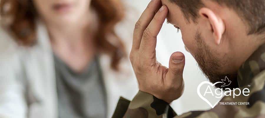 Person in camouflage jacket holding head with blurred female in background.