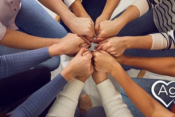 People sitting in a circle with hands joined in the center. Agape Treatment Center logo with heart outline.