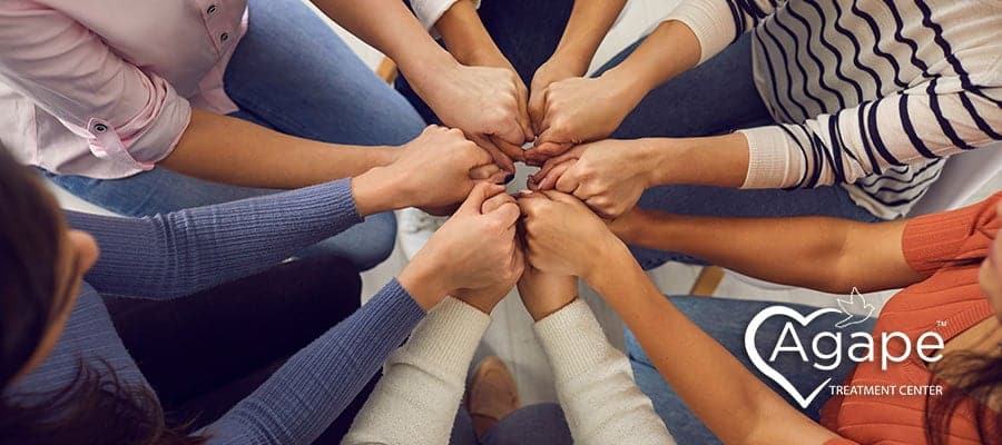 People sitting in a circle with hands joined in the center. Agape Treatment Center logo with heart outline.