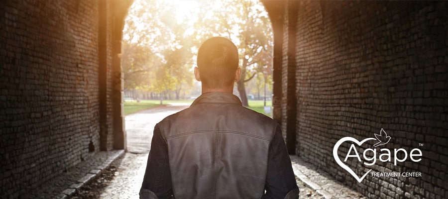 Person in a black jacket walking through a sunlit tunnel with Agape Treatment Center logo.