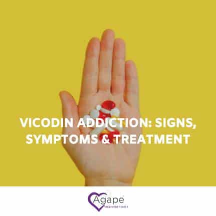 Hand holding pills against a yellow background with text Vicodin Addiction: Signs, Symptoms & Treatment.
