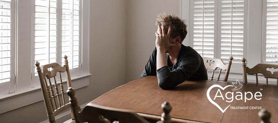 Person sitting at table with hand on face, near window shutters.