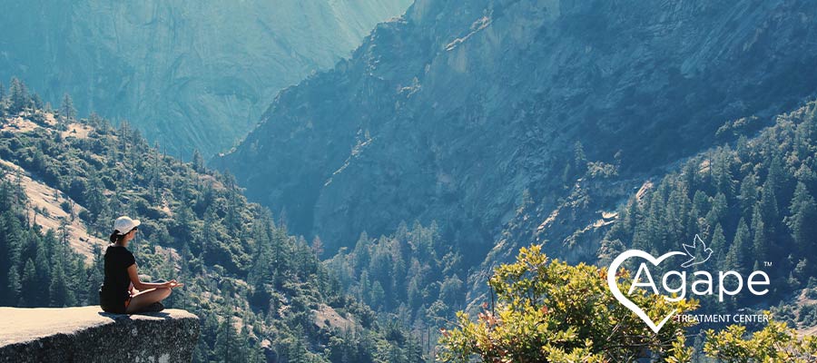 Person sitting on a rock edge in nature wearing a hat; Agape Treatment Center logo in white.