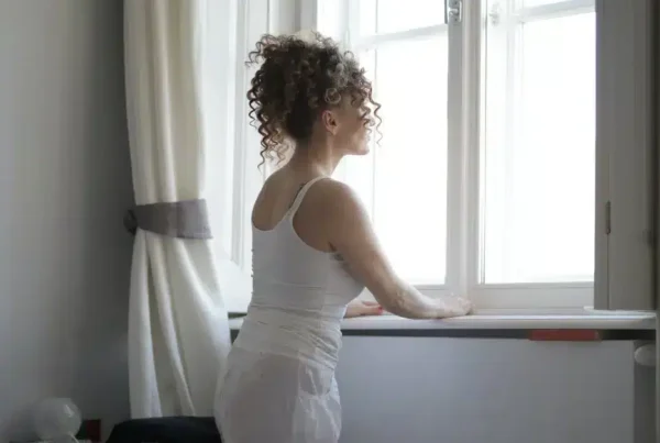 Person with curly hair wearing a white tank top looking out a window.