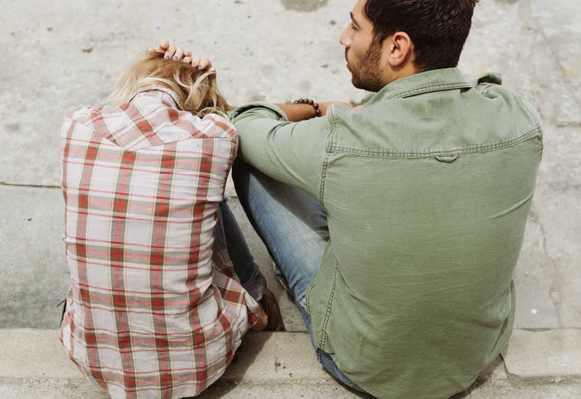 man and woman sitting together by a street while the woman has her head down