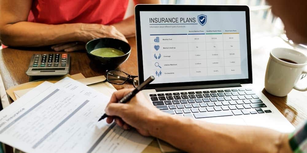 two people at a table with forms and a laptop open to insurance plan options