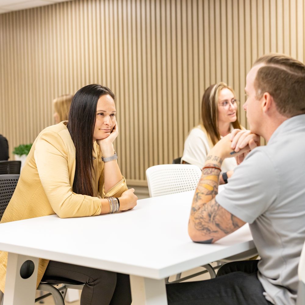 Person in tan jacket sitting at a table listening attentively.