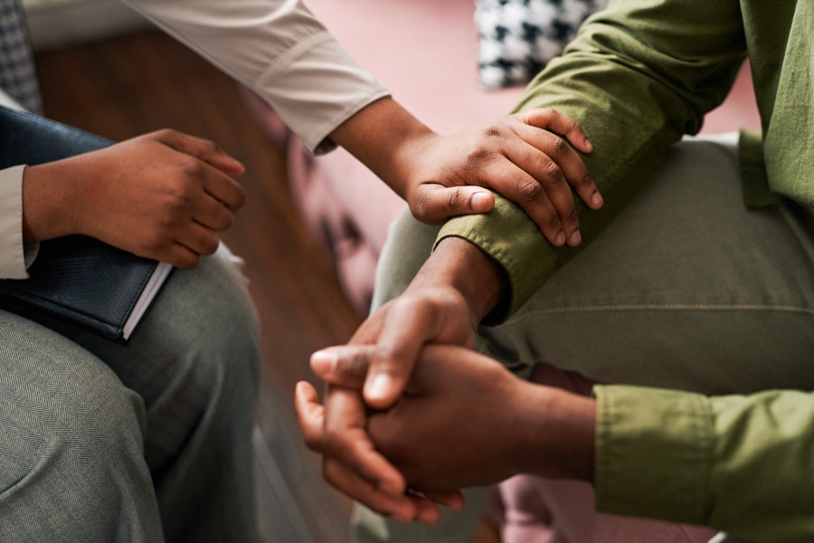 Two people sitting, one holding a notebook, the other wearing green, hands touching and supporting each other.