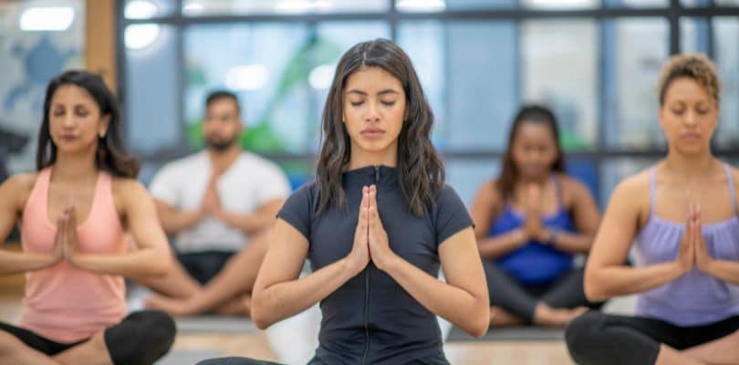 People sitting cross-legged in yoga poses, eyes closed, hands in prayer position.