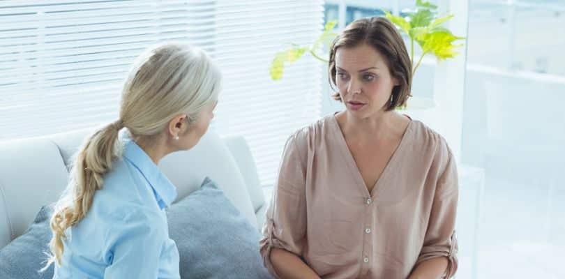 Two women sitting and talking in a bright room, one in a beige blouse, the other with a blue shirt.