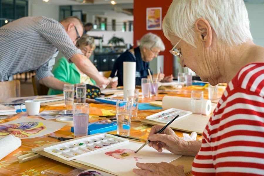 People painting at a table, elderly woman in red striped shirt painting with watercolors.