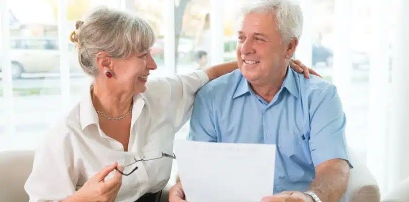 Elderly couple smiling, holding a document, woman in white blouse, man in blue shirt.