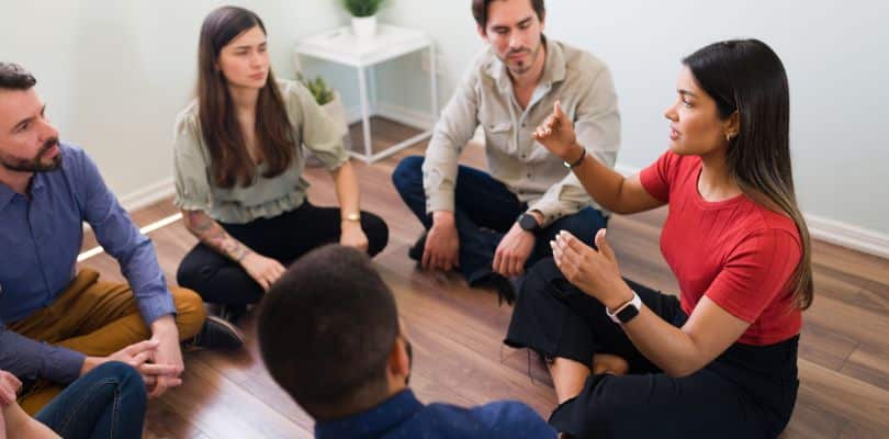 Person in a red shirt speaking to a seated group on a wooden floor.