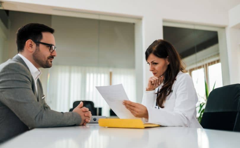 Person in white discussing papers with a seated man in a gray suit.