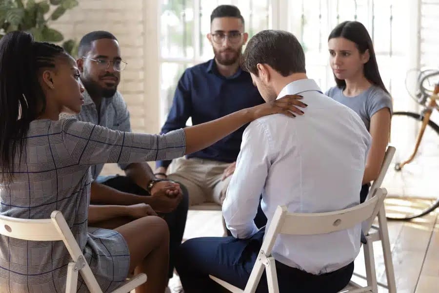 People sitting in a circle, one woman in checkered dress comforting a man in white shirt.