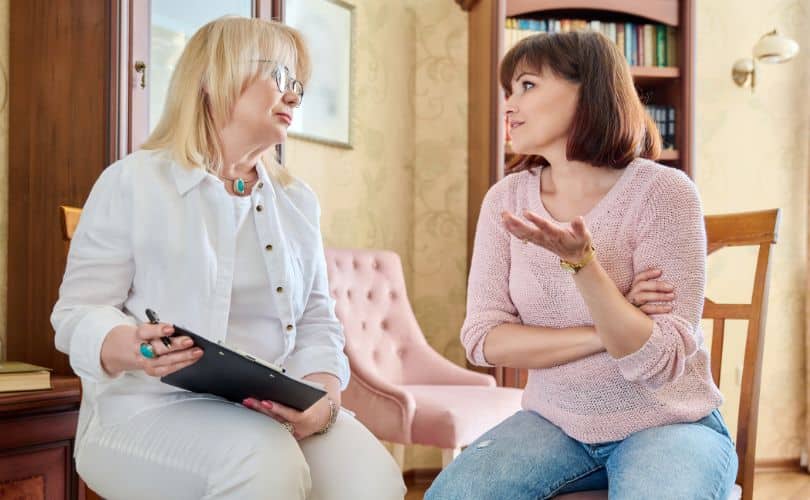 Two people sitting and talking, one in a white shirt with a notepad, the other in a pink sweater gesturing.