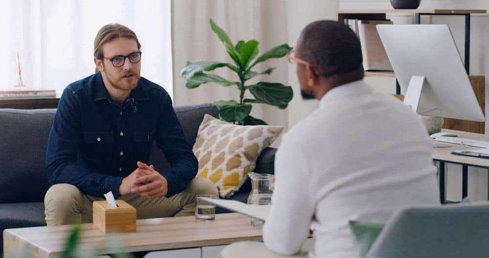 Person wearing glasses and denim shirt speaking with another person in a modern office setting.